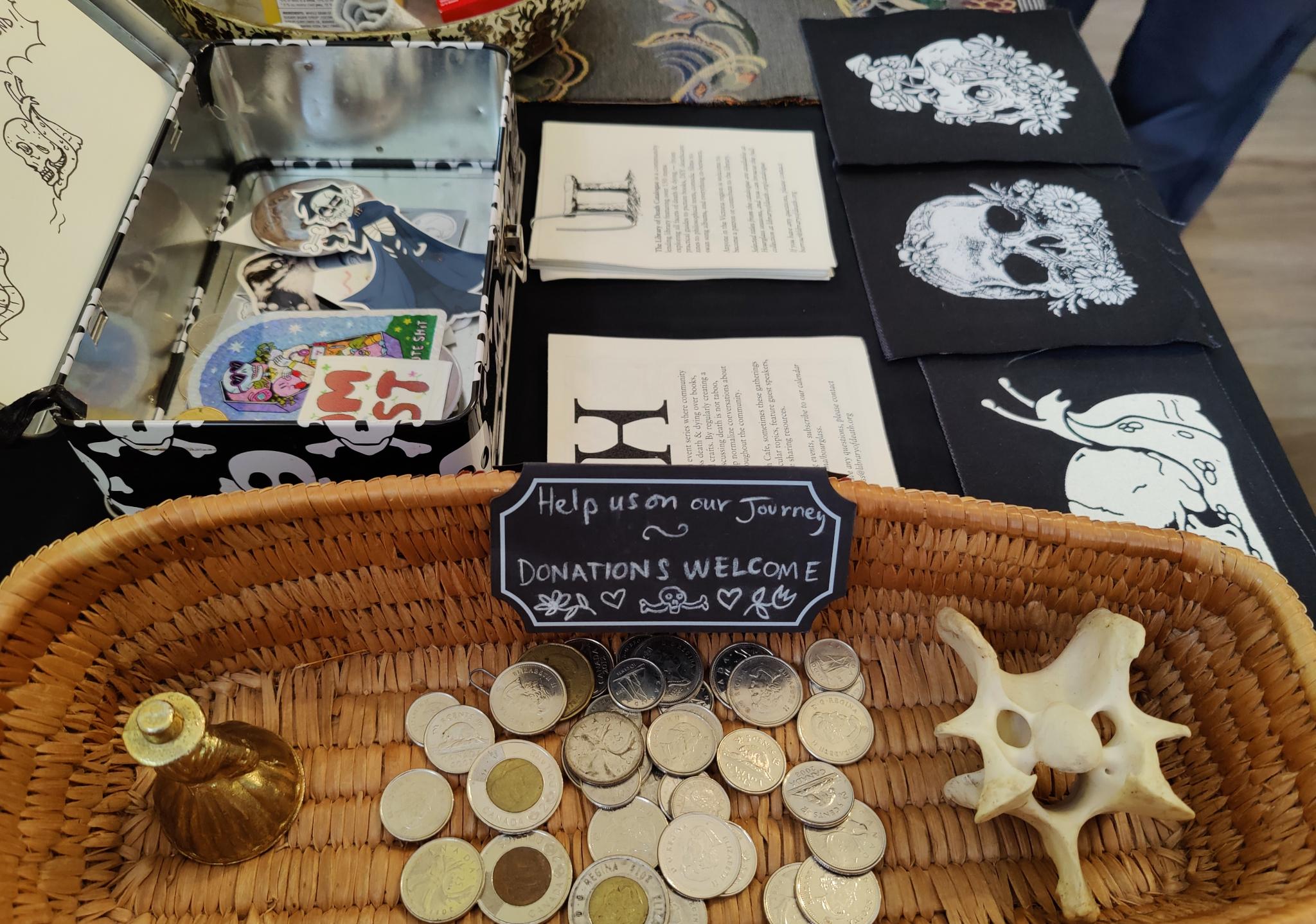 A donation basket with coins inside, along with a bell and a bone. Behind sits a small metal container with various death-themed stickers and patches.
